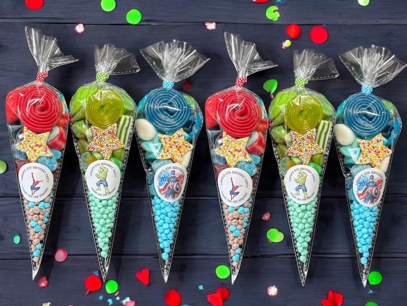 May include: Six cellophane cone-shaped bags filled with sweets. Each bag is tied at the top and features a different superhero sticker. The bags contain lollipops, star-shaped sprinkles, and other colourful treats. The bags are on a dark blue surface.