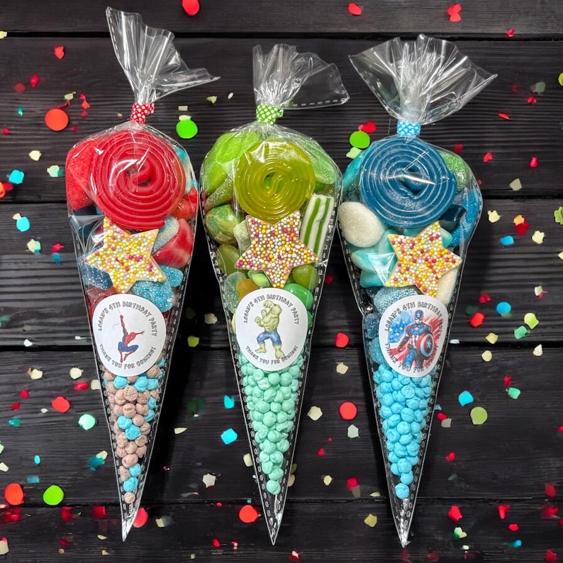 May include: Three cellophane cone-shaped bags filled with sweets. Each bag has a different superhero sticker and is tied with a ribbon. The bags contain lollipops, gummy sweets, and chocolate candies. The text on the sticker reads "4th Birthday Party".