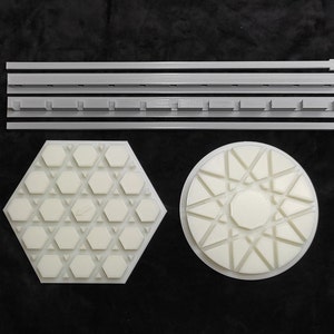 May include: Three white plastic pieces with geometric designs. The first piece is a hexagonal shape with a honeycomb pattern. The second piece is a round shape with a starburst pattern. The third piece is a long, rectangular piece with a series of holes.