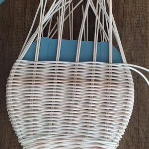 May include: A light cream-coloured, oval-shaped woven basket in the process of being made. Inside the basket is a rectangular block of blue foam, with many thin, light-coloured wooden sticks rising upwards. The basket sits on a dark wood surface.