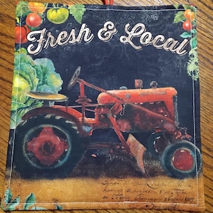 Puede incluir: Un agarrador de tela cuadrado con un lazo rojo para colgar. El diseño presenta un tractor rojo vintage sobre un fondo negro con las palabras "Fresh & Local" en escritura blanca. Verduras verdes y rojas bordean la parte superior.