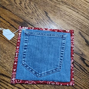 Puede incluir: Posavasos cuadrado con bolsillo de mezclilla y borde con estampado de bandana roja. El posavasos está hecho de mezclilla reciclada y presenta un bolsillo funcional. El borde rojo tiene un estampado de cachemira blanco. Una etiqueta blanca está adjunta.