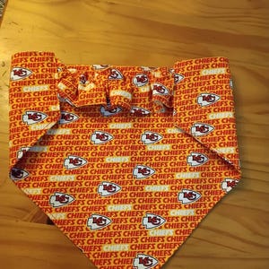 May include: A dog bandana with a red and white pattern, displaying the word "CHIEFS" and the team logo. It has a frilled top edge and a triangular shape.