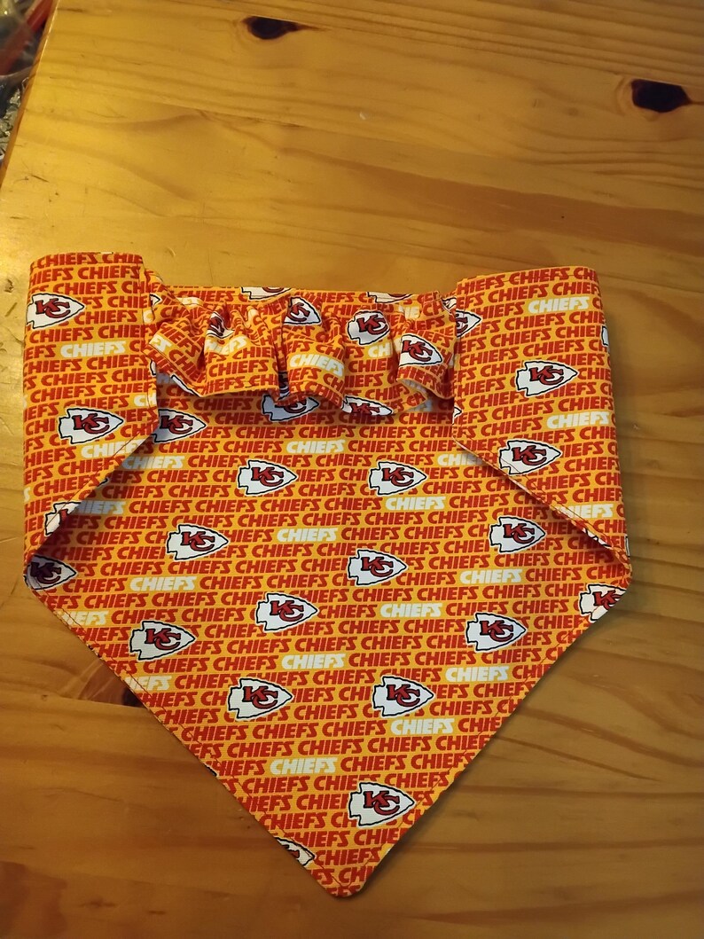 May include: A dog bandana with a red and white pattern, displaying the word "CHIEFS" and the team logo. It has a frilled top edge and a triangular shape.