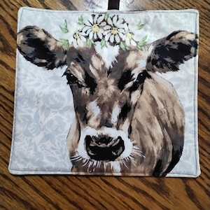 Op de afbeelding: Een bruine en witte koe met een krans van madeliefjes op haar hoofd. De koe kijkt recht in de camera. De afbeelding is op een witte achtergrond met een bloemenpatroon.