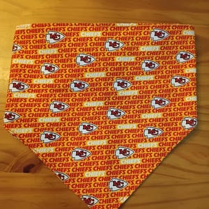 May include: A triangular pet bandana with a repeating pattern of the Kansas City Chiefs logo and the word "CHIEFS" in red, white, and gold. The bandana is made of fabric.