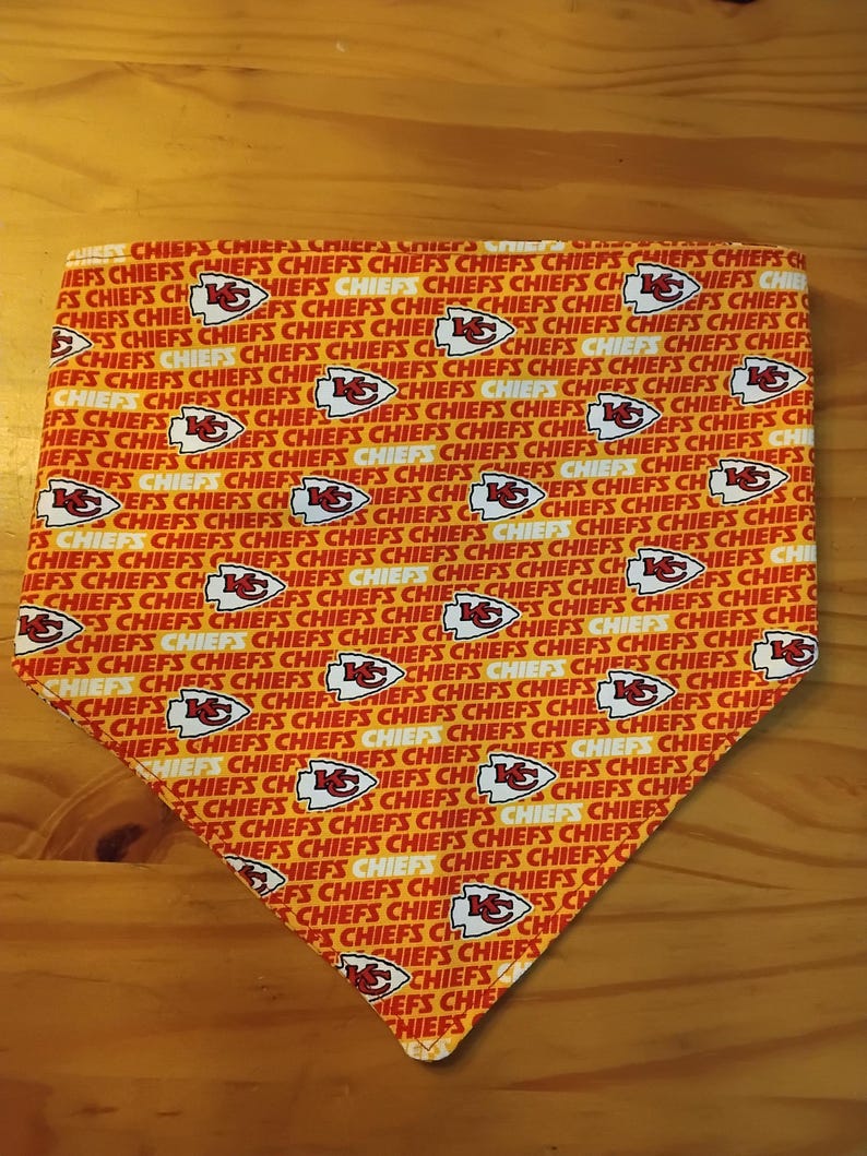 May include: A triangular pet bandana with a repeating pattern of the Kansas City Chiefs logo and the word "CHIEFS" in red, white, and gold. The bandana is made of fabric.