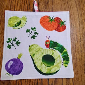 以下が含まれることがあります： 赤色の縁取りのある白いキッチンタオル。アボカドの上の緑色の毛虫、紫色の玉ねぎ、緑色のアボカドの半分、緑色の鳥、赤いトマト2つが描かれている。タオルには赤と白のチェック柄の吊り下げループが付いている。