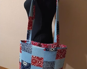 May include: A patchwork tote bag in blue, red, and white patterned fabrics. The bag has paisley, gingham, and solid colour designs. The straps are made of the same patchwork fabric. The bag is displayed on a mannequin.