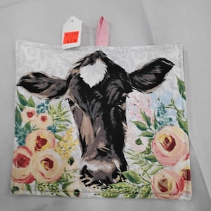 May include: A white cotton pot holder with a pink hanging loop. The pot holder features a colorful floral design with a cow's face in the center. The price tag reads "$2.99".
