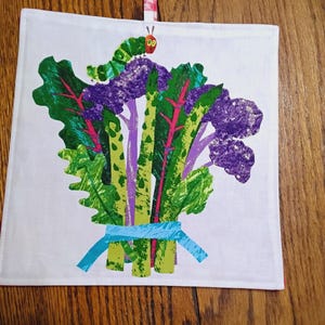 Może przedstawiać: Biały ręcznik kuchenny z kolorową ilustracją wiązki warzyw związanych niebieską wstążką. Zielona gąsienica z czerwonymi oczami znajduje się na szczycie warzyw.
