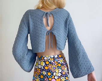 May include: A light blue knitted crop top with a tie back detail. The top has a diamond pattern and long sleeves. The model is wearing a colorful floral print skirt.