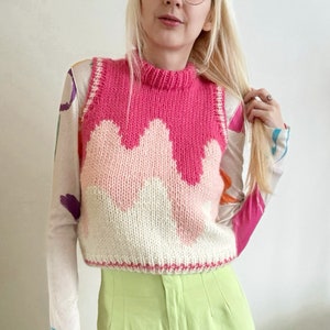 Può includere: Un gilet di maglia rosa e bianco con un motivo ondulato. Il gilet è senza maniche e ha un collo alto.