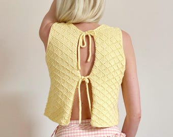 May include: A light yellow, sleeveless, open-back knit top. The top features a textured, diamond-patterned design and ties at the back. The bottom of the top is paired with a pink and white gingham skirt.