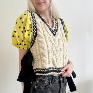May include: A cream-colored cable knit vest with black trim. The vest has a V-neck and is cropped. It is worn over a yellow and black polka dot blouse.