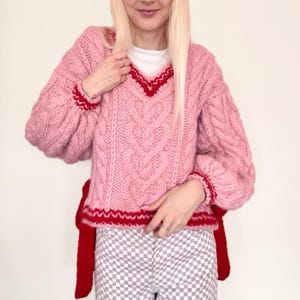 May include: A pink cable knit sweater with red trim and a V-neck. The sweater has a tie at the waist and is made of a soft, fluffy yarn.