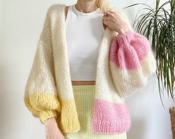 Può includere: Un cardigan di maglia bianco, giallo e rosa con maniche lunghe e una vestibilità ampia. Il cardigan ha una fascia di colore a contrasto sul fondo delle maniche e un collo a V.