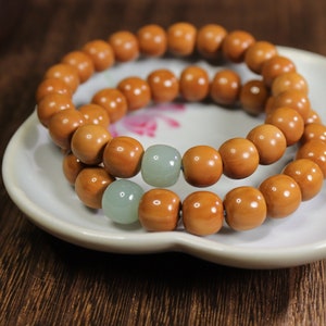 Pode incluir: Um bracelete de contas de madeira com duas contas de jade verde claro. O bracelete está em um prato de cerâmica branca.
