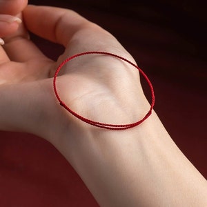 May include: A close-up of a red string bracelet resting on a person's palm. The bracelet is a simple, thin cord, tied in a loop. The background is a blurred, reddish-brown.