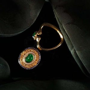 May include: Two pieces of jewelry: a gold ring with a heart-shaped design and a gold ring with a large oval green stone. The second ring has a gold filigree design with blue and white accent stones. Both rings are set against a dark background.