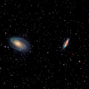 May include: A deep space image showcasing two galaxies against a backdrop of countless stars. The larger galaxy exhibits a spiral structure with hues of blue, yellow, and orange. The smaller galaxy is elongated, with red and blue tones.