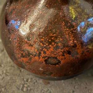 Könnte beinhalten: Eine kleine, runde Keramikvase mit einer dunkelbraunen und orangefarbenen, gesprenkelten Glasur. Die Vase hat eine glänzende Oberfläche und eine leicht unebene Oberfläche.