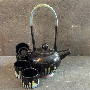 May include: A black ceramic teapot with a tall, arched handle and a matching set of three small cups. The teapot and cups have a glossy finish with yellow and blue vertical stripes. The teapot is approximately 10 inches tall.