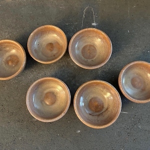 May include: Six small, brown ceramic bowls with a light brown glaze. The bowls are arranged in a semi-circle on a dark gray surface.