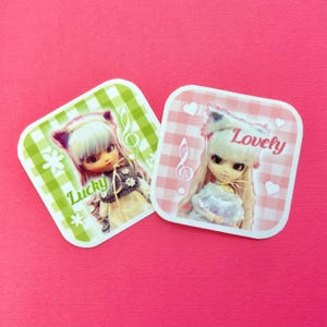 Puede incluir: Dos pegatinas cuadradas con ilustraciones de muñecas. Una tiene un fondo a cuadros verde y blanco con la palabra "Lucky" y una nota musical. La otra tiene un fondo a cuadros rosa y blanco con la palabra "Lovely" y una nota musical.