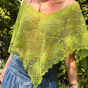 Puede incluir: Un poncho de ganchillo verde lima, de tejido abierto, con cuello en V. El poncho tiene un borde festoneado con detalles florales. La prenda se combina con una falda vaquera. El fondo es un exuberante follaje verde.