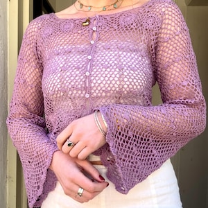 May include: A light purple crochet top with long, flared sleeves and a button-up front. The open weave design reveals a white underlayer. The top is paired with white bottoms. The top is a delicate, handmade garment.