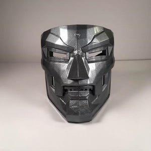 May include: A metallic gray mask with a geometric design, featuring rectangular eye and mouth openings. The mask has a textured surface and a distinct, angular shape, reminiscent of a superhero or villain's headgear.