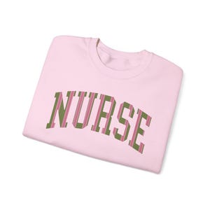 May include: A pale pink sweatshirt featuring the word "NURSE" in a green and pink striped font. The folded garment displays a crew neck design. The soft fabric is visible.