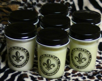 Puede incluir: Ocho pequeñas velas blancas en frascos de metal negro con un diseño de flor de lis y el texto "Carnival Candle Company".