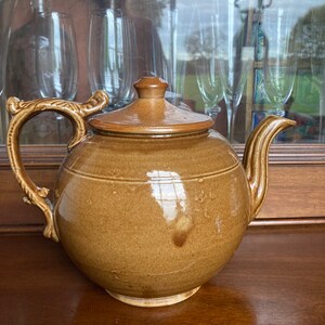 May include: A brown ceramic teapot with a lid and a curved handle. The teapot has a glazed finish and a slightly rounded body.