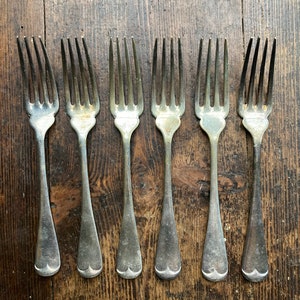 May include: Six silver forks with ornate handles, arranged in a row on a wooden surface.