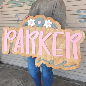 May include: Wooden sign with the name "Parker" in pink letters. The sign has a scalloped edge and is decorated with flowers in white and gray. The word "rae" is below the name in gray. A decorative piece for a child's room.