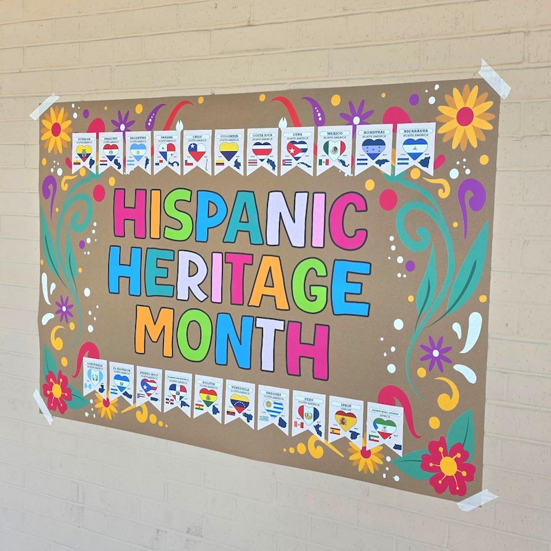 Op de afbeelding: Een groot bruin affiche met de woorden "Hispanic Heritage Month" in kleurrijke letters. De poster is versierd met bloemmotieven en kleine vlaggen die verschillende Spaanse landen vertegenwoordigen. De poster is vastgeplakt aan een bakstenen muur.