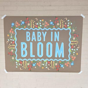 Op de afbeelding: Een bruine poster met de woorden "BABY IN BLOOM" in lichtblauw, omlijst door een lichtblauwe rand. De rand is versierd met kleurrijke bloemen en groen, waaronder rode, oranje, blauwe en gele bloemen. De poster is met tape aan een bakstenen muur bevestigd.