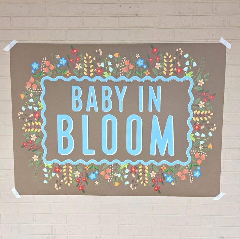 Op de afbeelding: Een bruine poster met de woorden "BABY IN BLOOM" in lichtblauw, omlijst door een lichtblauwe rand. De rand is versierd met kleurrijke bloemen en groen, waaronder rode, oranje, blauwe en gele bloemen. De poster is met tape aan een bakstenen muur bevestigd.