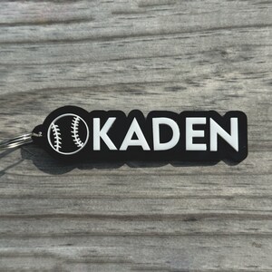 May include: Black and white baseball keychain with the name "KADEN" in white lettering.