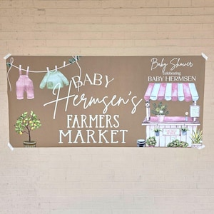 Op de afbeelding: Een bruine banner met de woorden "Baby Hermsen's Farmers Market" in witte letters. De banner bevat illustraties van babykleding, een citroenboom en een marktkraam met een roze en wit gestreepte luifel. De woorden "Baby Shower Celebrating Baby Hermsen" staan ook op de banner.