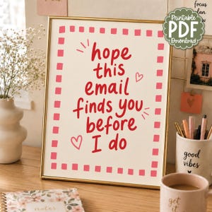 Può includere: Una stampa artistica rettangolare con cornice dorata, sfondo color crema, bordo a scacchi rossi e il testo rosso "hope this email finds you before I do", accompagnato da due piccole illustrazioni di cuori rossi e segni decorativi.