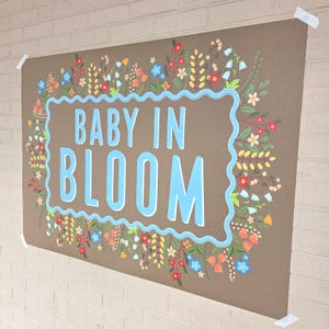 May include: A large brown poster with the words "BABY IN BLOOM" in light blue, surrounded by a decorative border and floral illustrations in various colors. The poster is taped to a white brick wall.