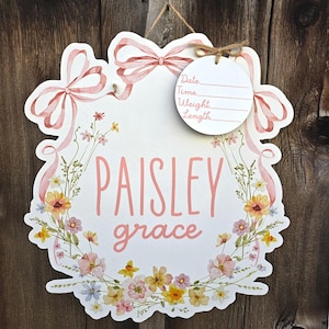 May include: A white, floral-themed wooden sign with the name "Paisley Grace" in pink script. The sign features a wreath of flowers and ribbons, with a small tag for recording birth details. A twine loop is attached for hanging.