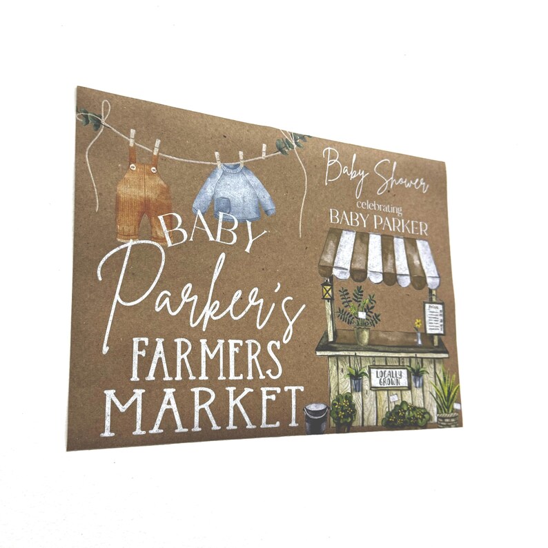 May include: A brown card with the text "Baby Parker's Farmers Market" in white script. The card features illustrations of baby clothes hanging on a washing line and a farmers market stall. The words "Baby Shower celebrating Baby Parker" are also present.