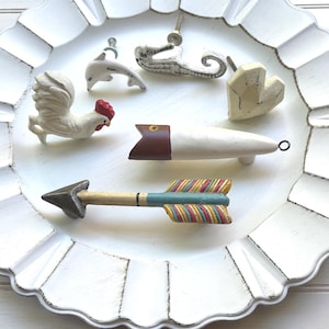 May include: A collection of decorative drawer pulls and knobs in various shapes, including a rooster, dolphin, alligator, heart, and arrow. The pulls are white, with red, brown, and colorful accents. The items are arranged on a white, scalloped-edge tray.