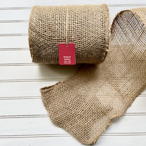 May include: A roll of natural burlap ribbon with a red tag. The burlap is a light brown color and has a woven texture. The tag reads "BURLAP Arpillera 14cm x 9m".
