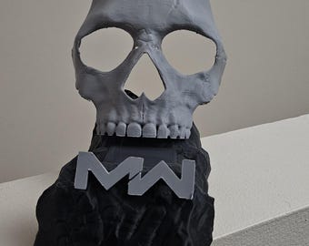 Könnte beinhalten: Eine graue Totenkopfskulptur auf einem strukturierten schwarzen Sockel. Der Totenkopf hat hohle Augenhöhlen und eine Nasenhöhle. Eine graue Plakette mit den Buchstaben MW ist an der Vorderseite des schwarzen Sockels befestigt, der eine raue Oberfläche hat.