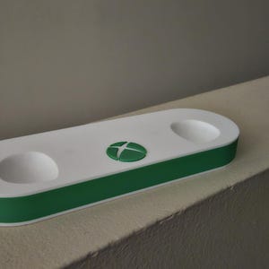 K&ouml;nnte beinhalten: Eine wei&szlig;-gr&uuml;ne Controller-Ladestation in Pillenform mit zwei runden, vertieften Aussparungen f&uuml;r Controller und einem mittigen gr&uuml;nen runden Logo mit einem wei&szlig;en, vierquadrantigen Symbol auf der Oberseite.
